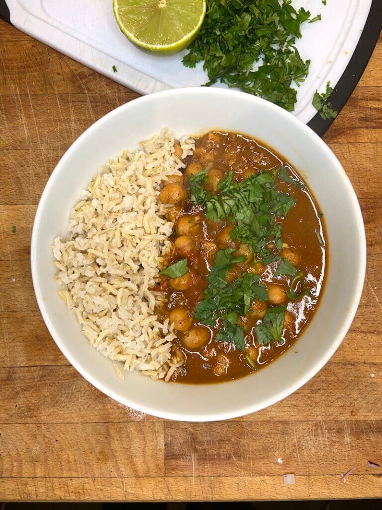 bowl of vegan coconut chickpea curry with coriander and brown rice, with half a lime and coriander on the back 