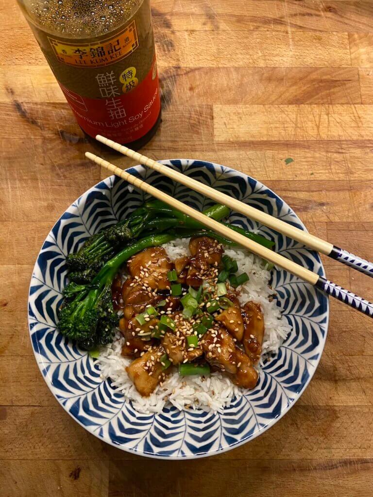 chopsticks and white and blue bowl of chicken teriyaki topped with toasted sesame seeds and spring onions, accompanied by tenderstem broccoli and boiled white rice