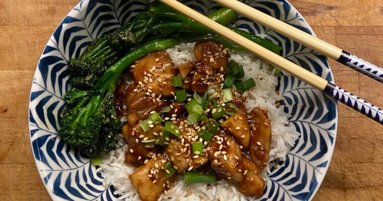 white and blue bowl with chicken teriyaki on a bed of rice and covered with spring onions and toasted sesame seeds with a side of stir fry broccoli