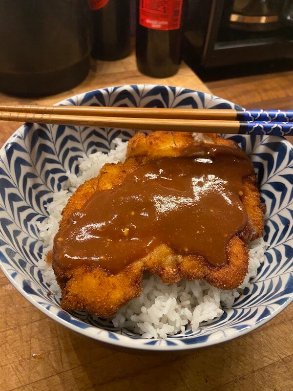 white and blue decorated ramen bowl with boiled rice, a slice of deep fried chicken katsu topped with katsu sauce and two decorated chopsticks laying on top