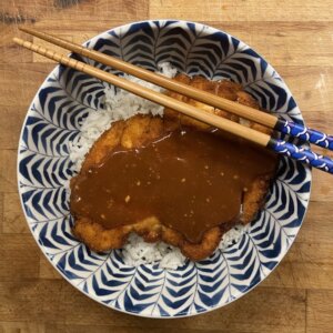 a white and blue decorated bowl with a bed of rice, a fried chicken breast coated in breadcrumbs, topped with a Katsu sauce set on a wooden cutting board and with two decorated chopsticks on top