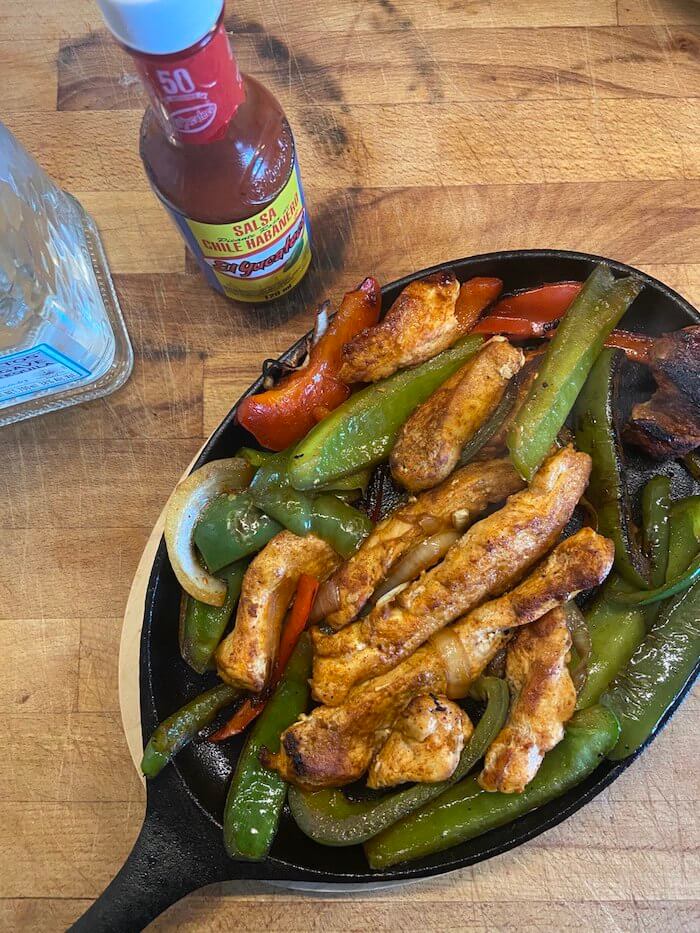 sizzling fajitas pan with a mix of chicken, red and green peppers and onion photographed next to hot sauce and a bottle of tequila