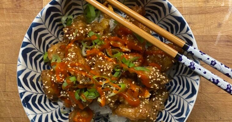 A decorated bowl of sticky lemon chicken over a bed of rice, garnished with spring onions, sesame seeds and sriracha and with a pair of decorated chopsticks on top