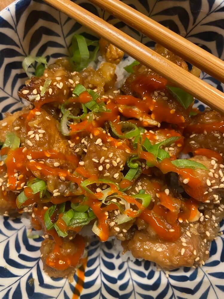 close up of a bowl of sticky lemon chicken topped with sesame seeds, sriracha and green onions over a bed of boiled rice