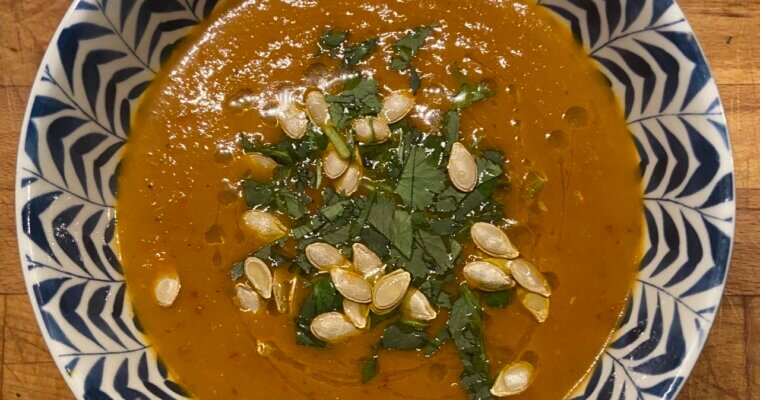 Bowl of spiced pumpkin soup topped with roasted pumpkin seeds, chopped fresh coriander and olive oil. Sitting on a wooden butting board.